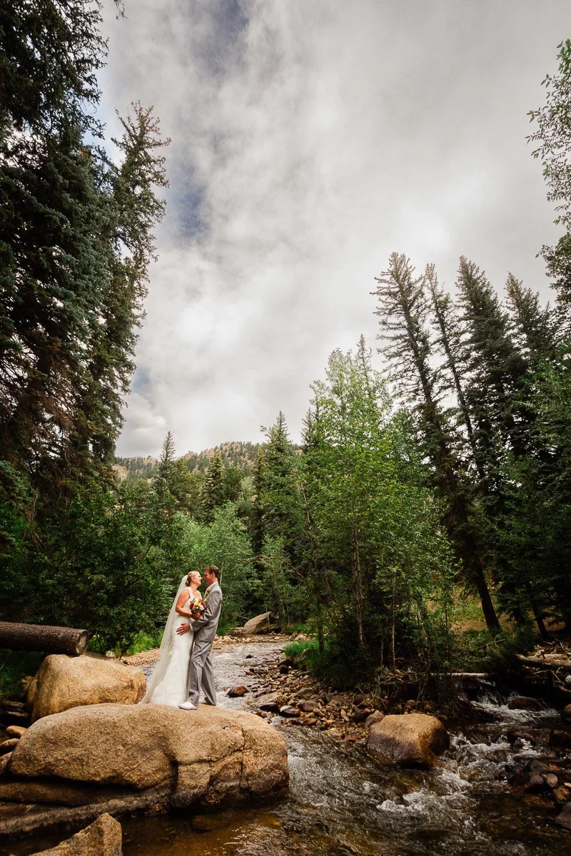 A bride and groom embrace on a large rock beside a flowing river in a serene forest. Towering pine trees surround them under a cloudy sky, evoking a peaceful and romantic atmosphere.