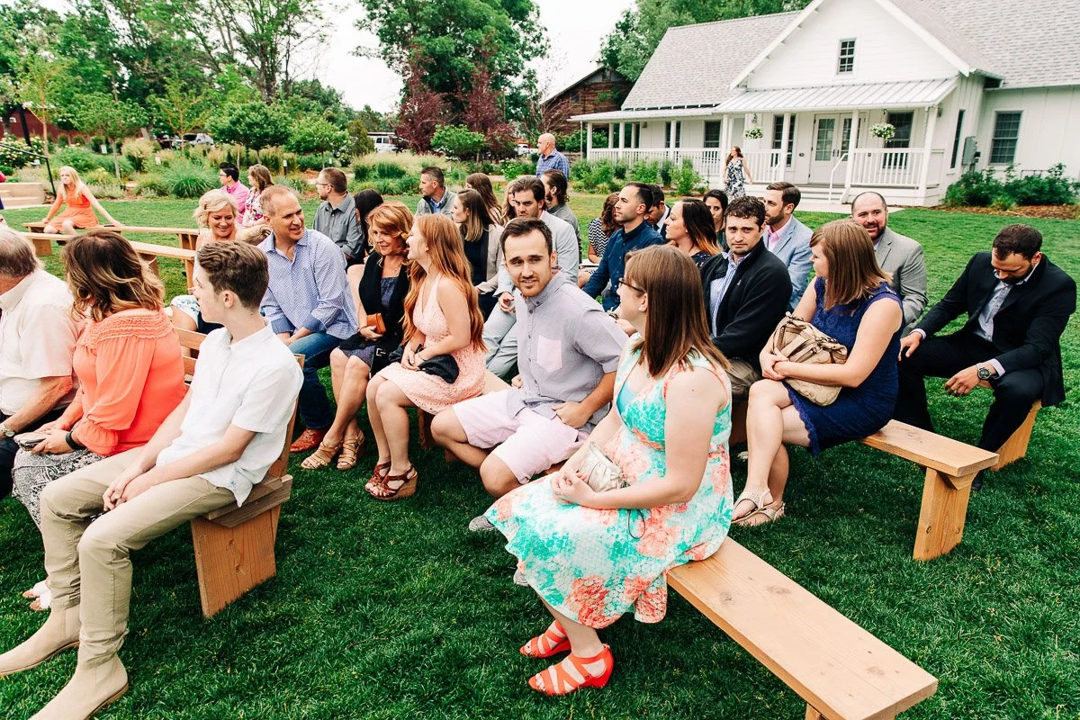 A group of people seated on wooden benches outdoors, surrounded by greenery and a white house. They're dressed in semi-formal attire, engaged in conversation, creating a joyful and relaxed atmosphere.