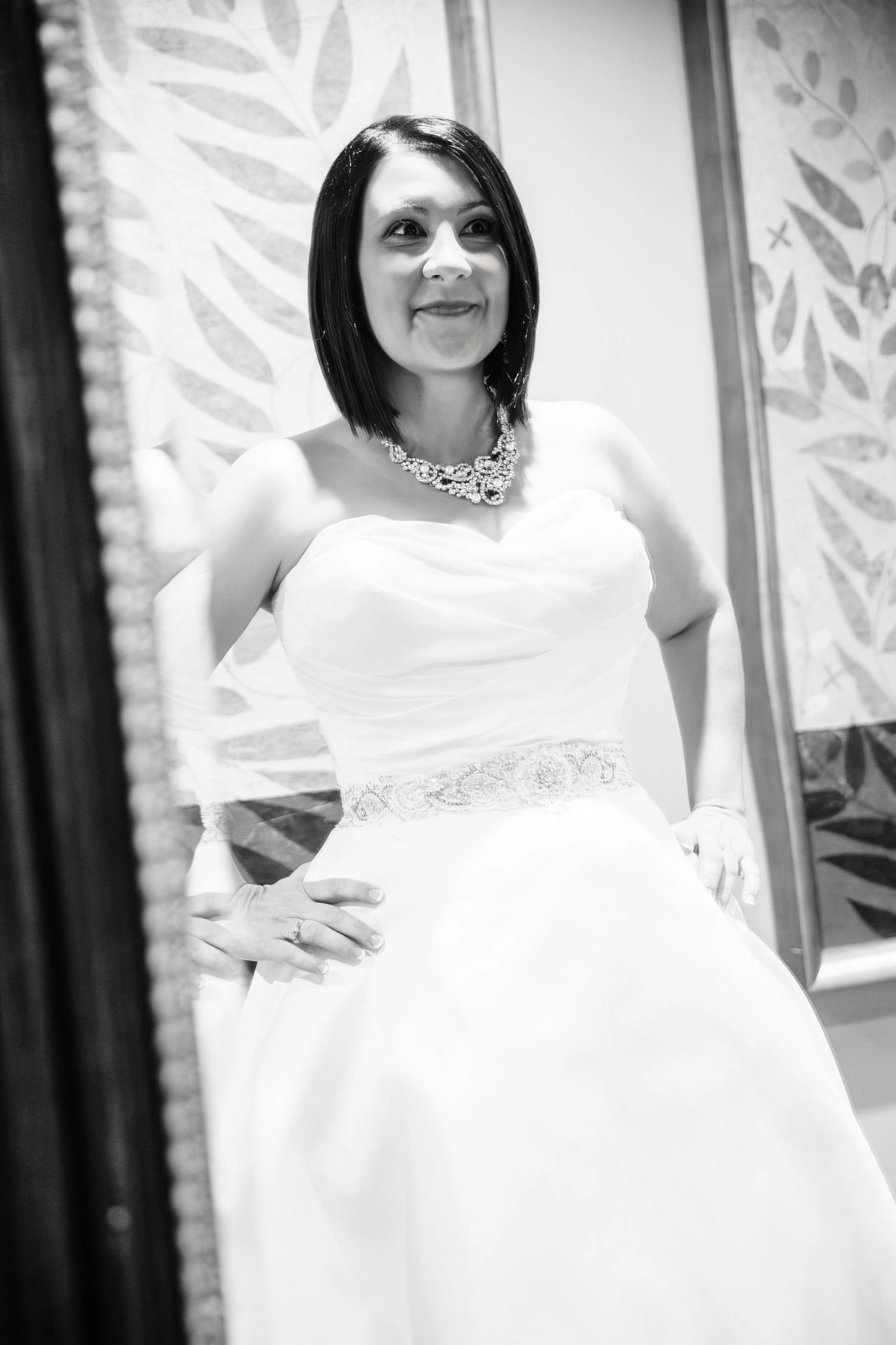 A woman stands confidently in a strapless wedding dress, hands on hips. The black-and-white photo captures her joyful expression and elegant necklace.
