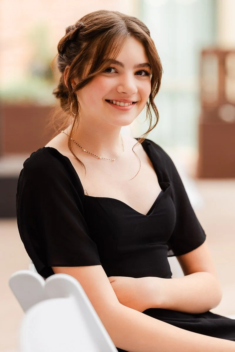A young woman with brown hair, wearing a black dress and necklace, smiles warmly. Her hair is styled in an updo. The background is softly blurred.