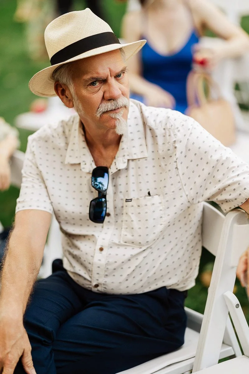 Elderly man with a white goatee and straw hat sits in a garden setting, wearing a patterned shirt and sunglasses. He looks thoughtful and relaxed.