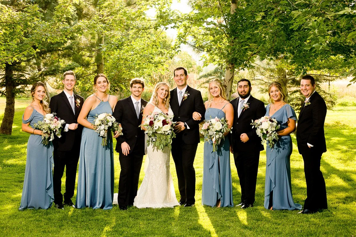 A wedding party poses outdoors on a sunny day. The bride in a white gown and groom in a black suit stand center, surrounded by bridesmaids in blue dresses and groomsmen in black suits. They're all smiling under leafy trees, holding bouquets.