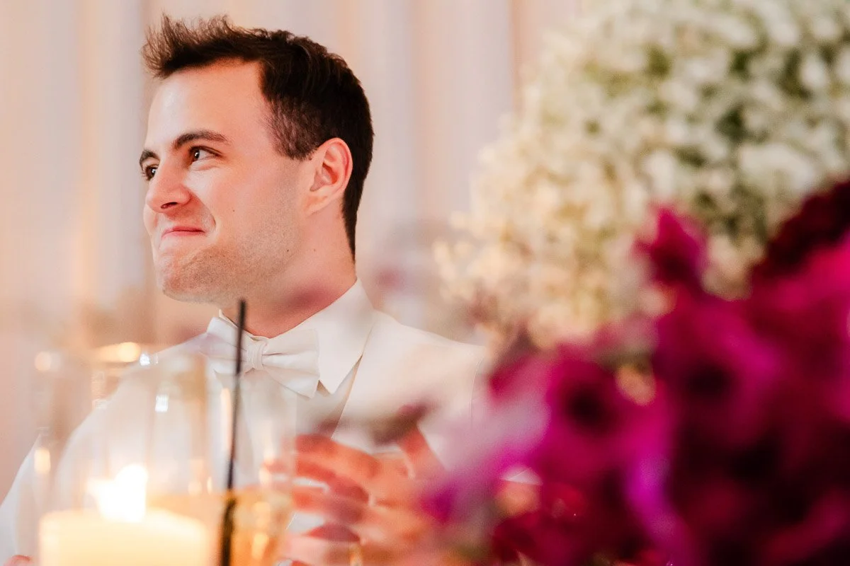 A smiling man in a white suit sits at a table adorned with candles and vibrant flowers. The scene is warm and joyful, suggesting a celebratory event.