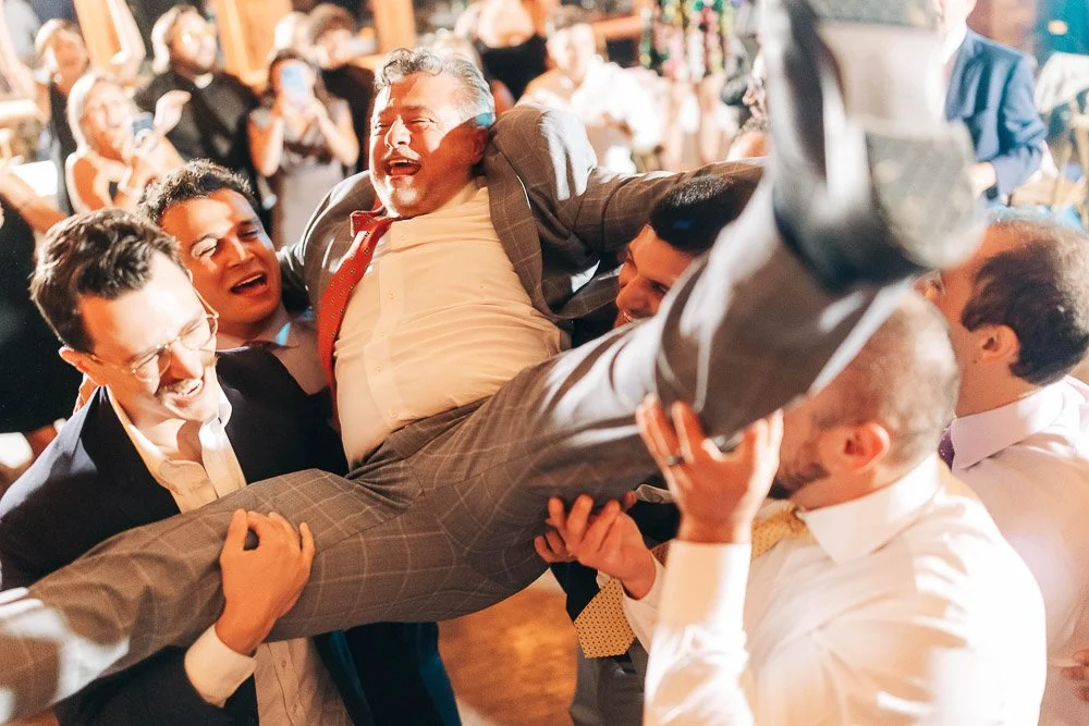 A group of laughing men in suits joyously lift an older man in a plaid suit during a lively celebration, creating a cheerful and festive atmosphere.