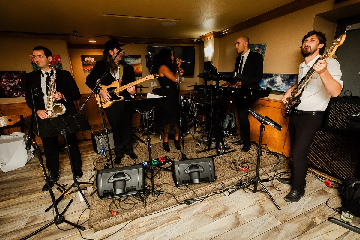 A five-member band performs energetically indoors. Musicians play saxophone, guitar, keyboard, and bass. Dim lighting creates an intimate atmosphere.