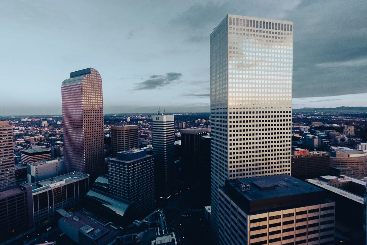 Aerial view of a Denver cityscape at dusk, featuring tall skyscrapers with reflective glass. The sky is overcast, creating a calm, subdued atmosphere.
