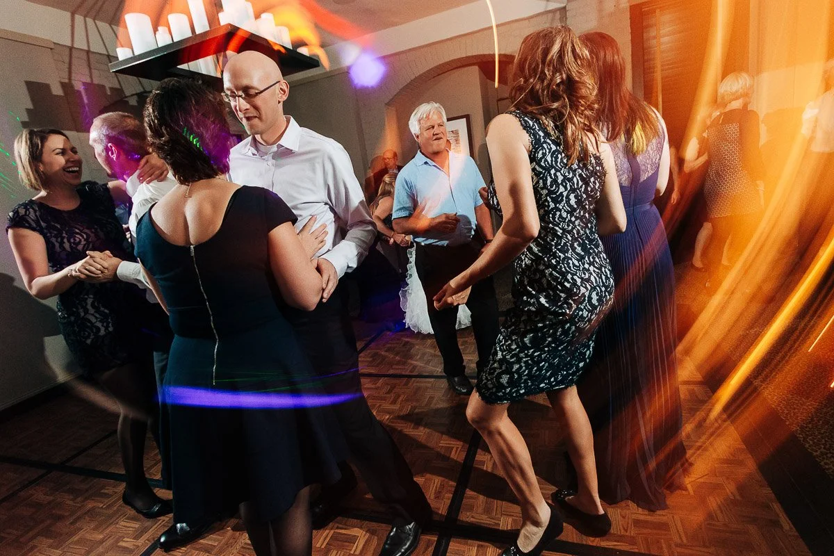 A lively dance party scene with people in formal attire, smiling and enjoying themselves on a wooden floor. Warm, colorful light trails add vibrancy.