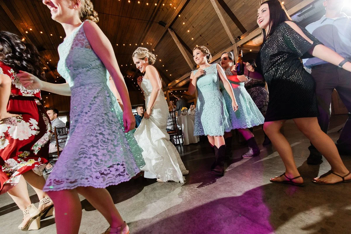 A group of people joyfully dancing at a wedding, with women in light blue dresses and a bride in white. Warm, festive lights hang overhead.