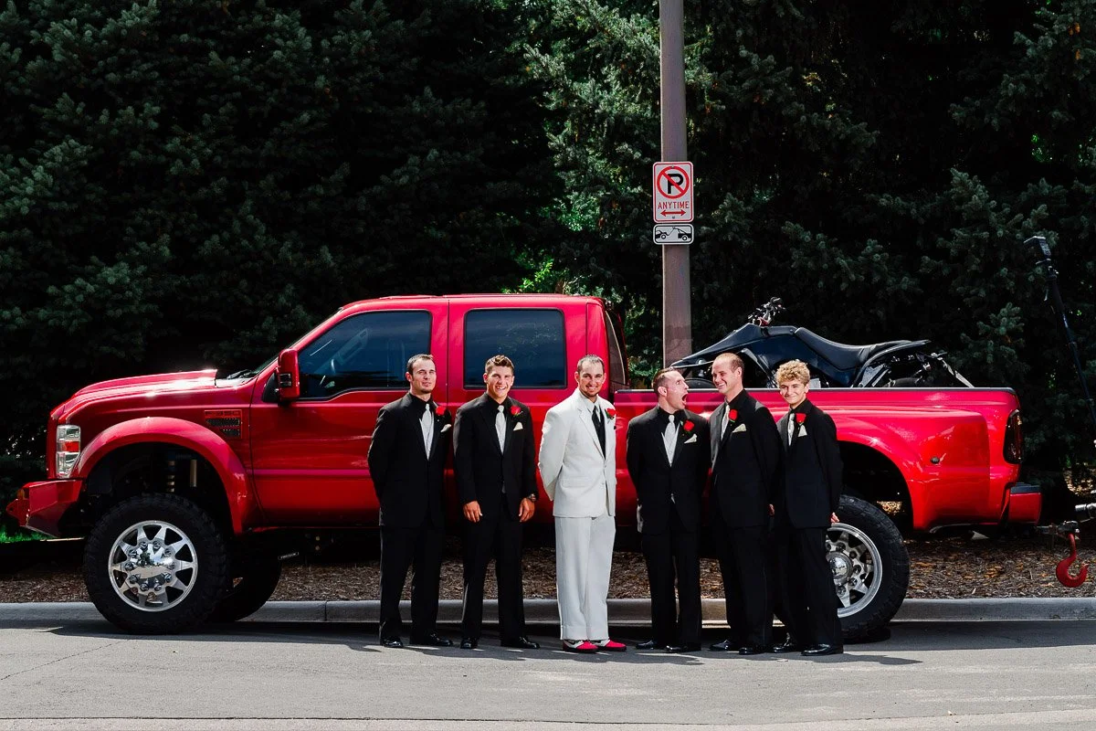 A group of six men in formal suits, one in a white tuxedo, stand beside a large red pickup truck with an ATV in the back, against a backdrop of tall trees.