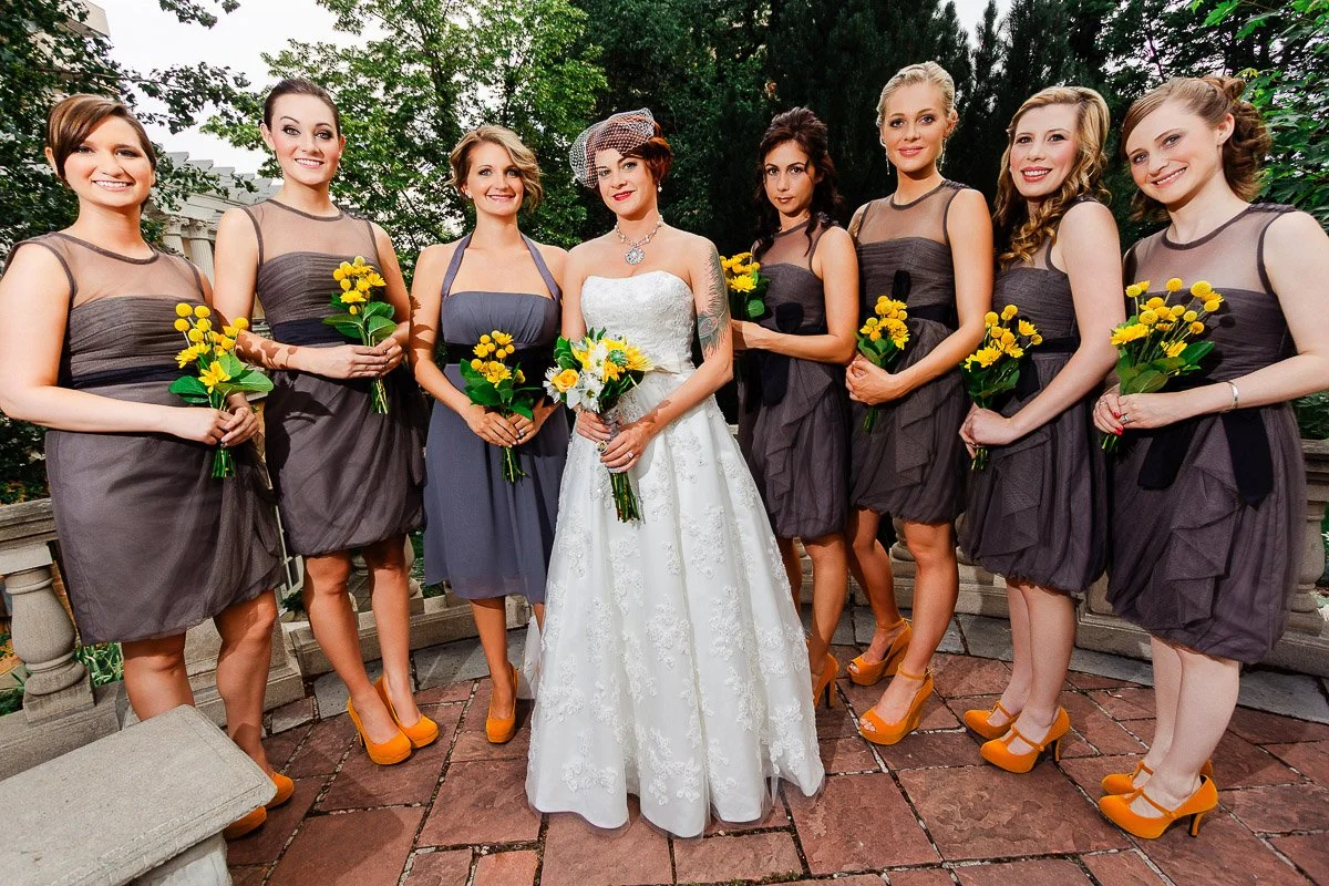 A bride in a white gown stands with seven bridesmaids in grey dresses holding yellow bouquets. The group is outdoors, exuding joy and celebration.