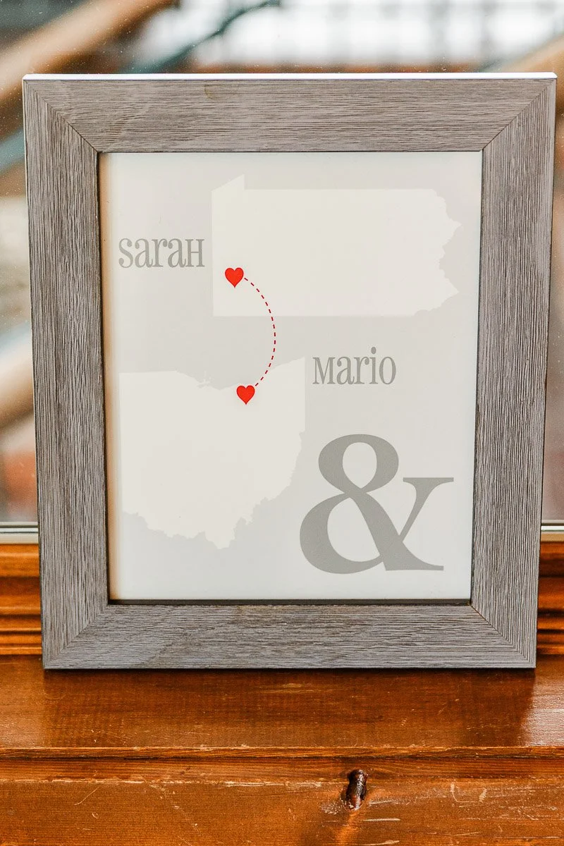Framed artwork with a map of two states connected by dotted line and hearts. Names "Sarah" and "Mario" are beside each state with an ampersand.