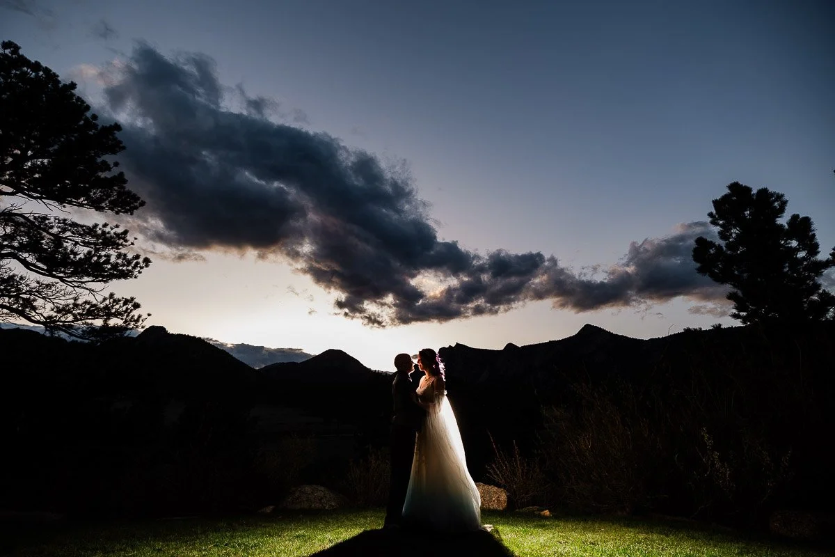Silhouetted bride and groom embrace under a dramatic evening sky during a Black Canyon wedding with billowing clouds, surrounded by mountains and trees, creating a serene and romantic atmosphere.
