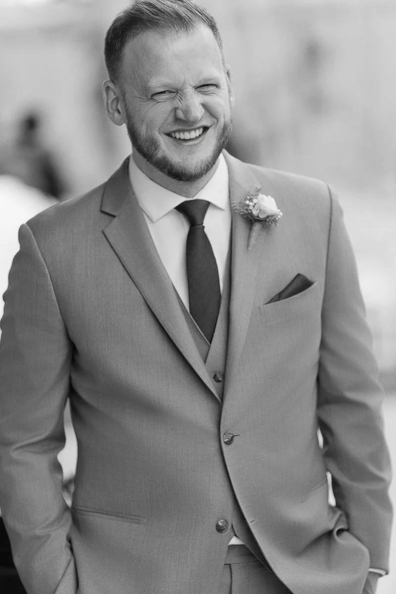 Man in a light suit with a boutonniere, smiling and squinting, hands in pockets. The background is blurred, emphasizing a joyful, candid moment.
