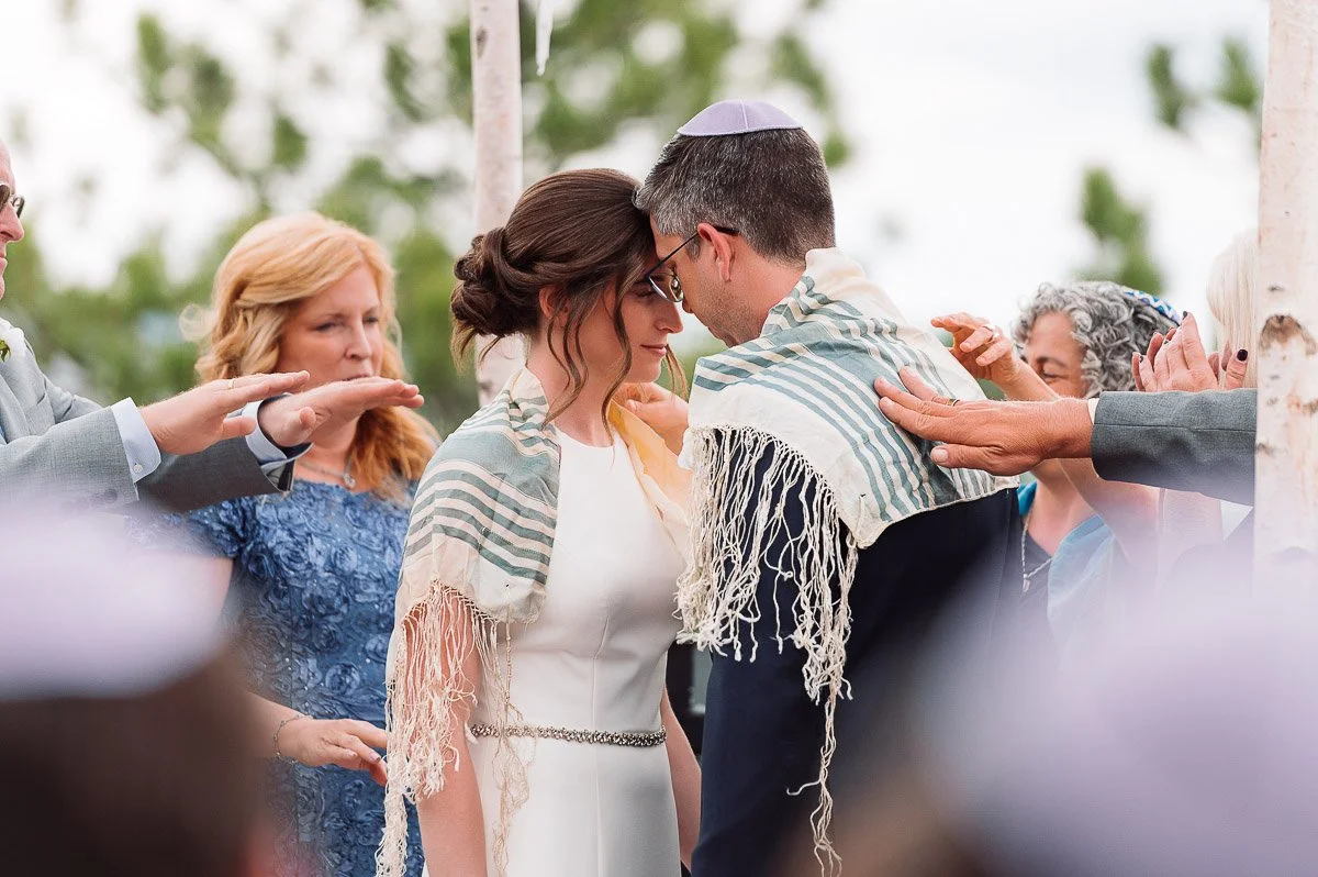 Bride and groom in a Jewish wedding ceremony, draped in prayer shawls under a chuppah. Embracing with closed eyes, surrounded by family with hands extended in blessing.