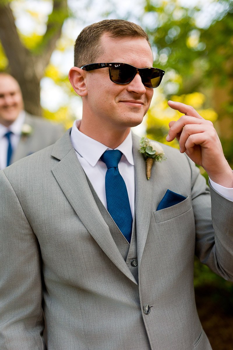 Man in a gray suit and blue tie adjusts his sunglasses outdoors. A boutonniere is pinned to his lapel, and another person is blurred in the background.