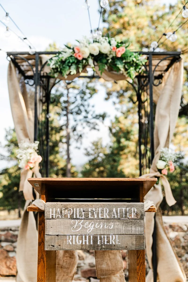 Rustic wedding altar outdoors, draped with burlap and adorned with a floral arrangement of roses and greenery. A wooden sign reads: "Happily Ever After Begins Right Here."