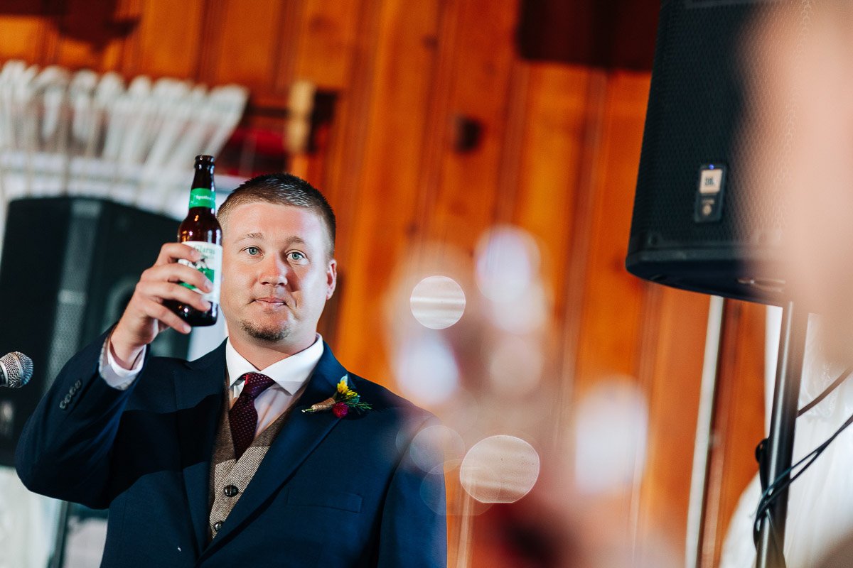 A man in a suit raises a beer bottle in a toast, standing in a warmly lit wooden room. The scene conveys celebration and camaraderie.