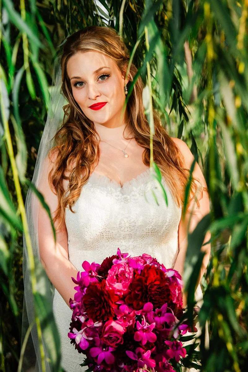 A bride stands amid green foliage, wearing a white lace dress and holding a vibrant bouquet of red and pink flowers. The scene is serene and joyful at a Lakewood Country Club wedding.