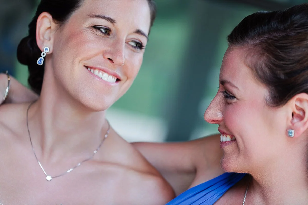 Two women smiling warmly at each other, one in a strapless dress, the other in blue, with jewelry, conveying happiness and friendship.