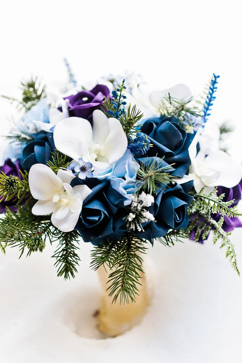 A vibrant bouquet with blue, purple, and white flowers, accented by green pine sprigs, set against a snowy background, evoking a winter theme.