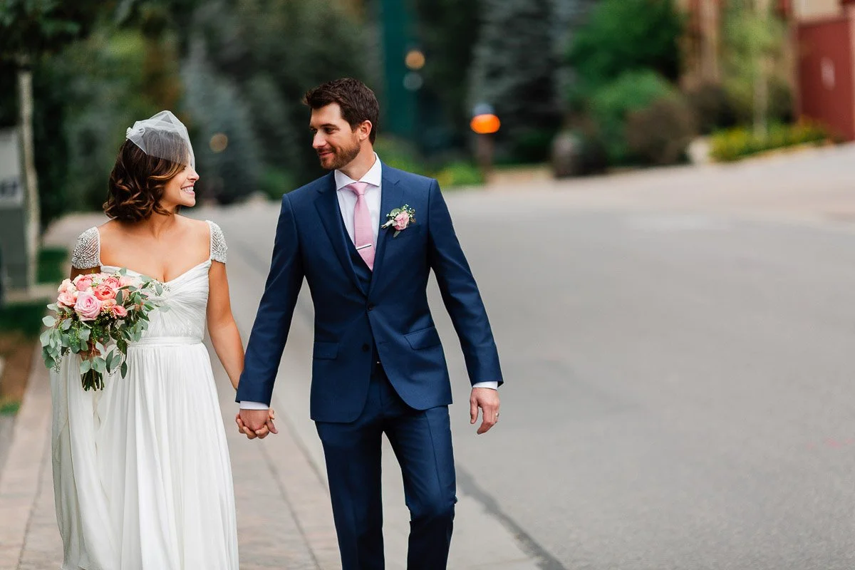 A bride in a white gown holds pink flowers, walking hand in hand with a groom in a blue suit on a tree-lined street, both smiling lovingly.