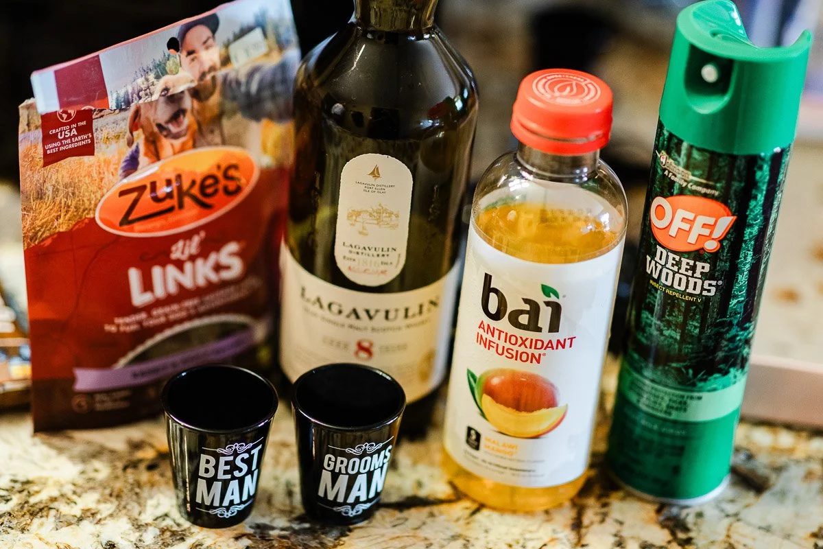 A collection of items on a counter: Zuke's pet treats, Lagavulin whisky, Bai drink, OFF! insect repellent, and two shot glasses labeled "Best Man" and "Grooms Man."