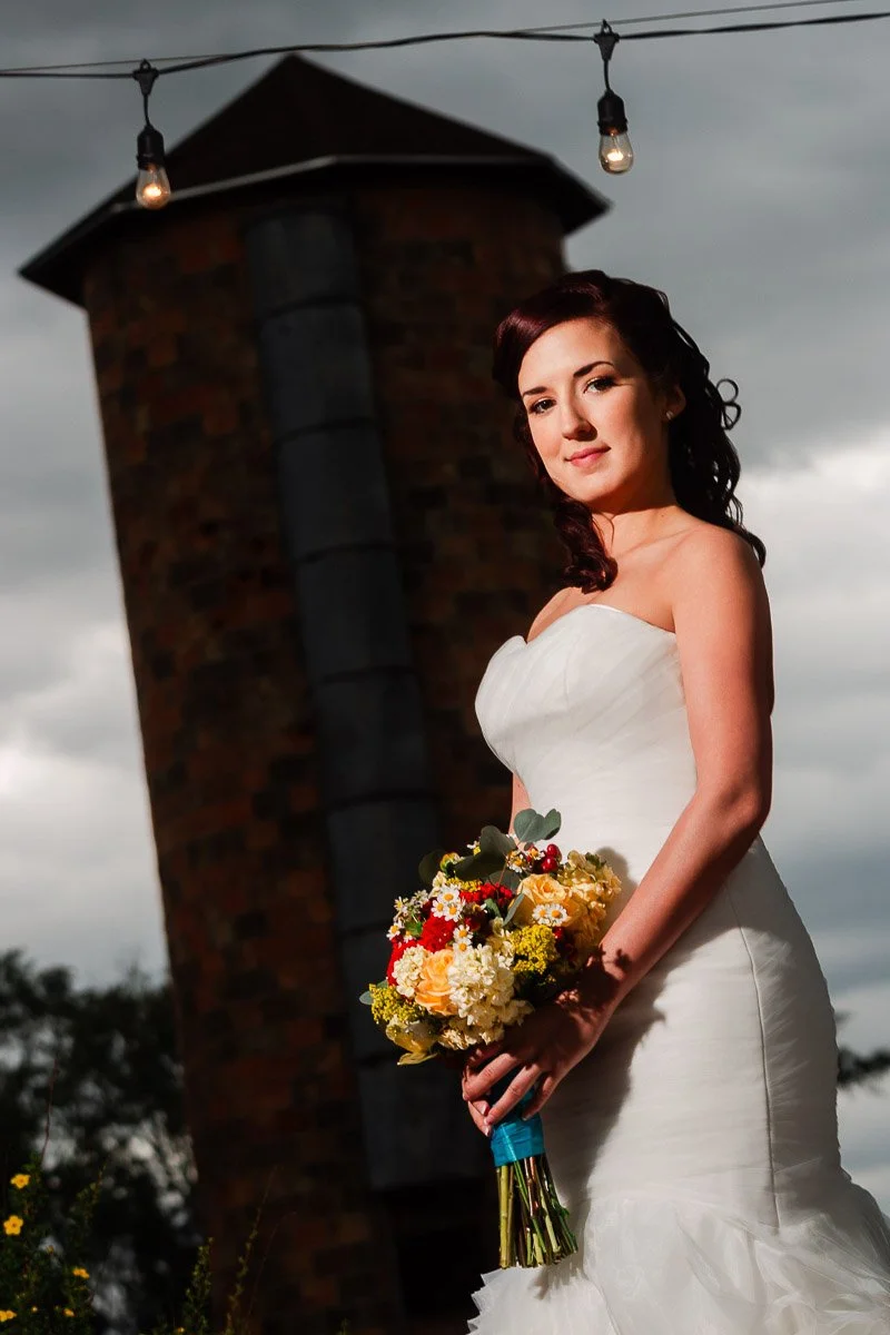 Bride in elegant white gown holding colorful bouquet stands confidently against a backdrop of a rustic brick tower and string lights under moody skies.