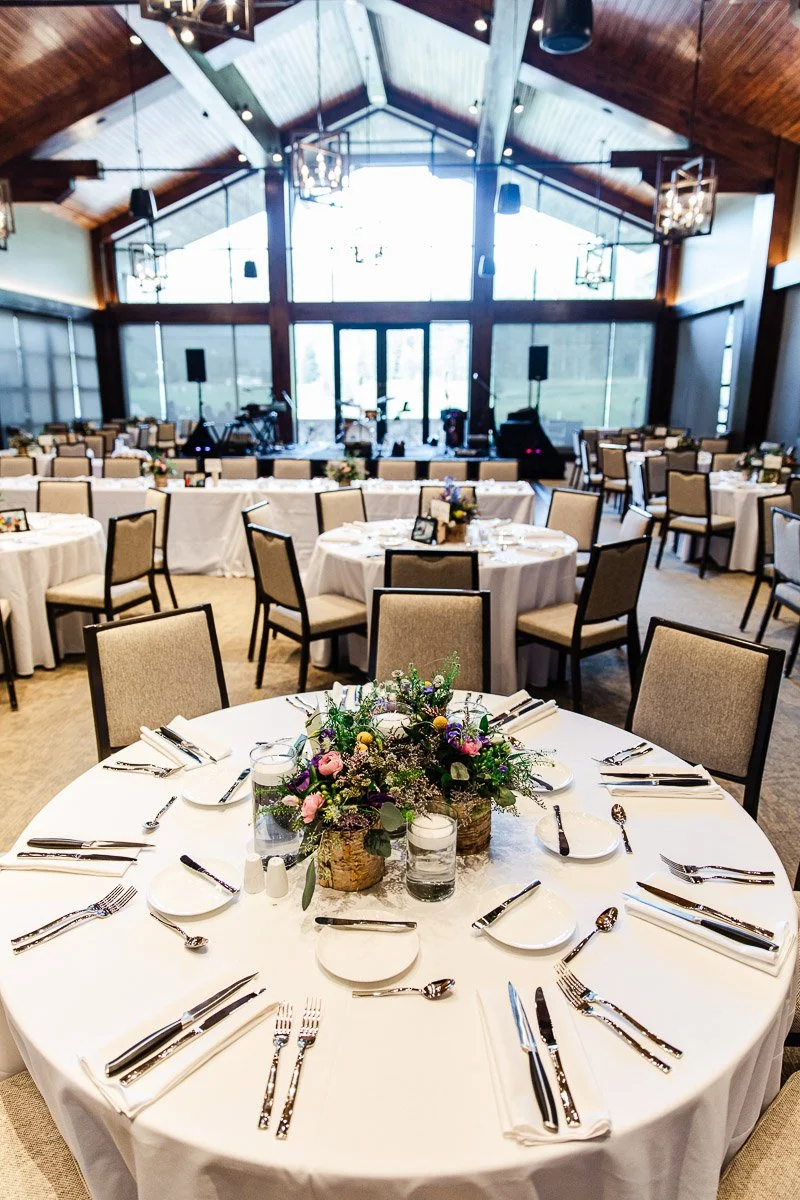 Elegant banquet hall with round tables set for an event. Tables have white cloths, floral centerpieces, and arranged silverware, under a wood-beamed ceiling.