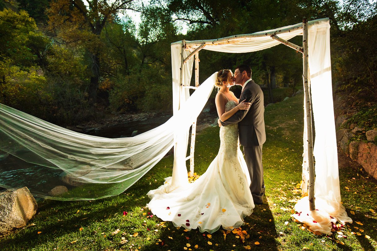 A bride and groom kiss under a rustic archway lit with soft sunlight, surrounded by greenery, with the bride's flowing veil billowing gently.