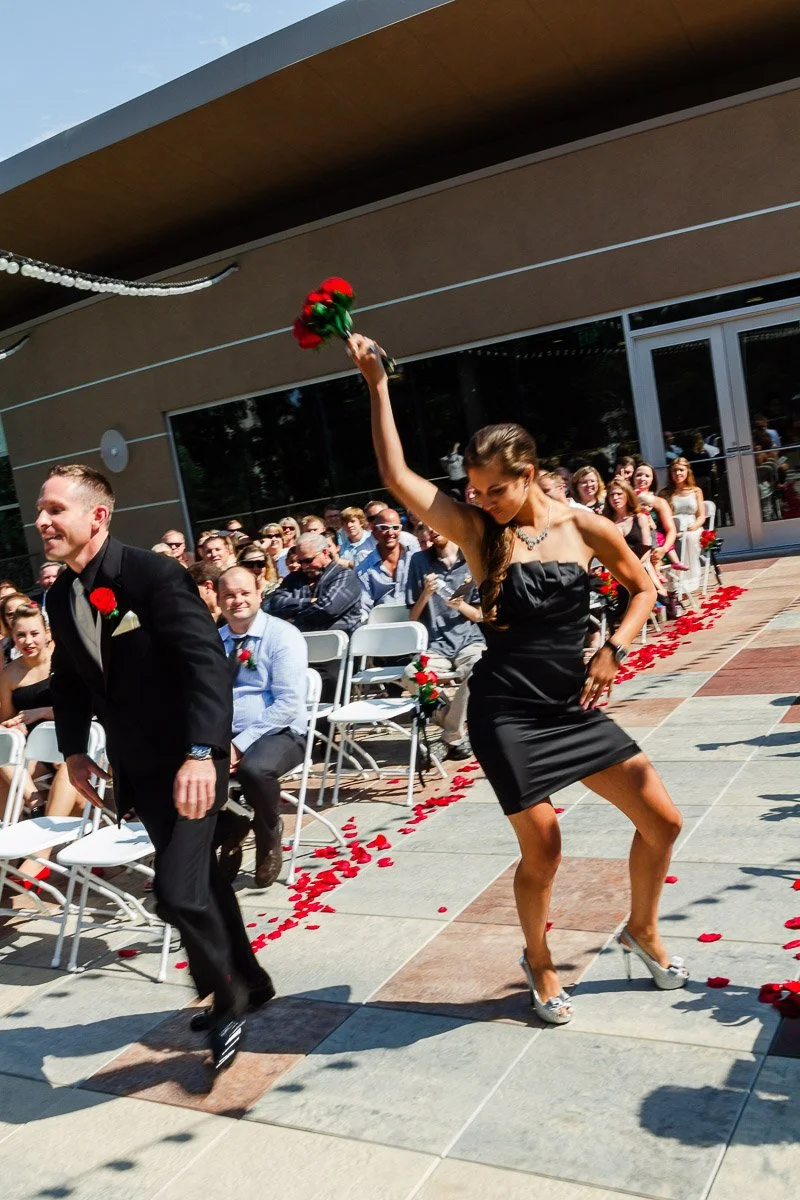 A joyful wedding scene with a woman in a black dress raising a red bouquet, dancing down an aisle strewn with rose petals, surrounded by seated, smiling guests.