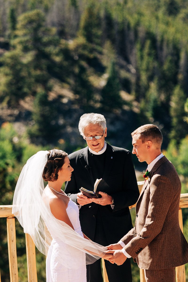 A bride and groom hold hands during an outdoor wedding ceremony. The officiant reads from a book. Forested mountains are in the background.