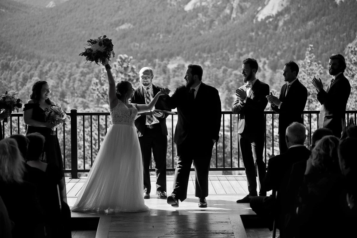 A bride and groom joyfully raise hands in a victory pose at a mountain-view wedding ceremony. Guests and attendants in formal wear applaud them.