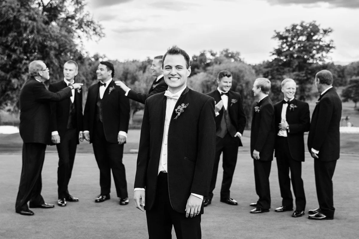 Smiling groom in a suit stands in the foreground. Groomsmen in tuxedos chat and adjust ties in the background. Outdoor setting, joyful atmosphere.