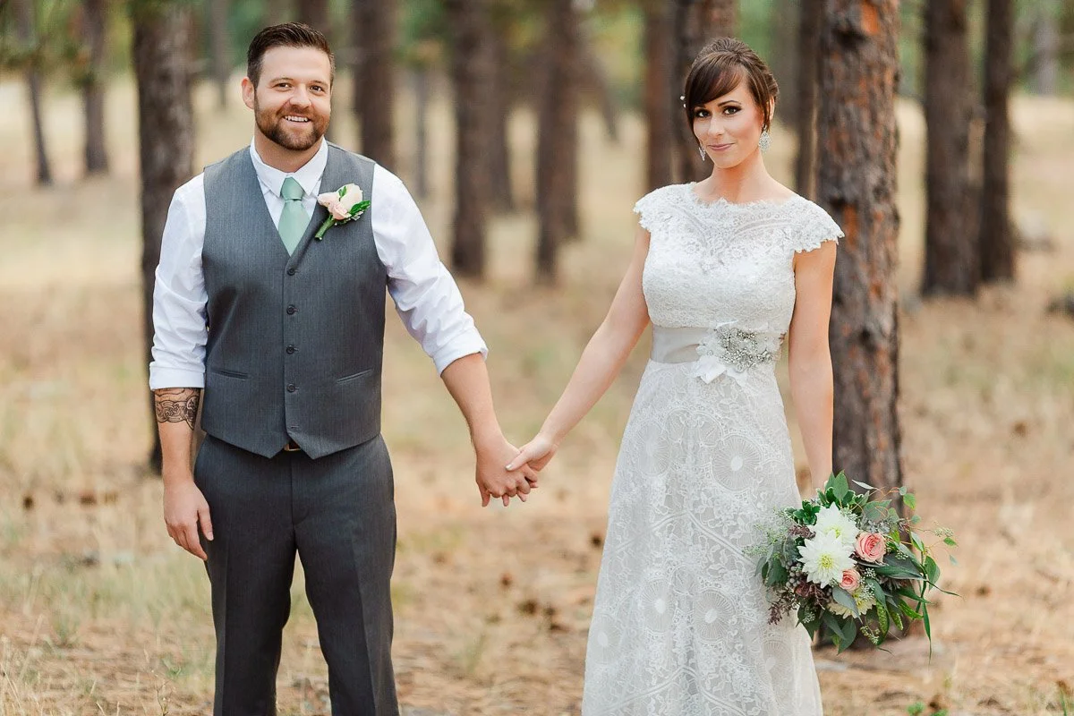 A couple in a forest, with a smiling man in a gray vest holding hands with a smiling woman in a lace wedding dress, holding a bouquet.