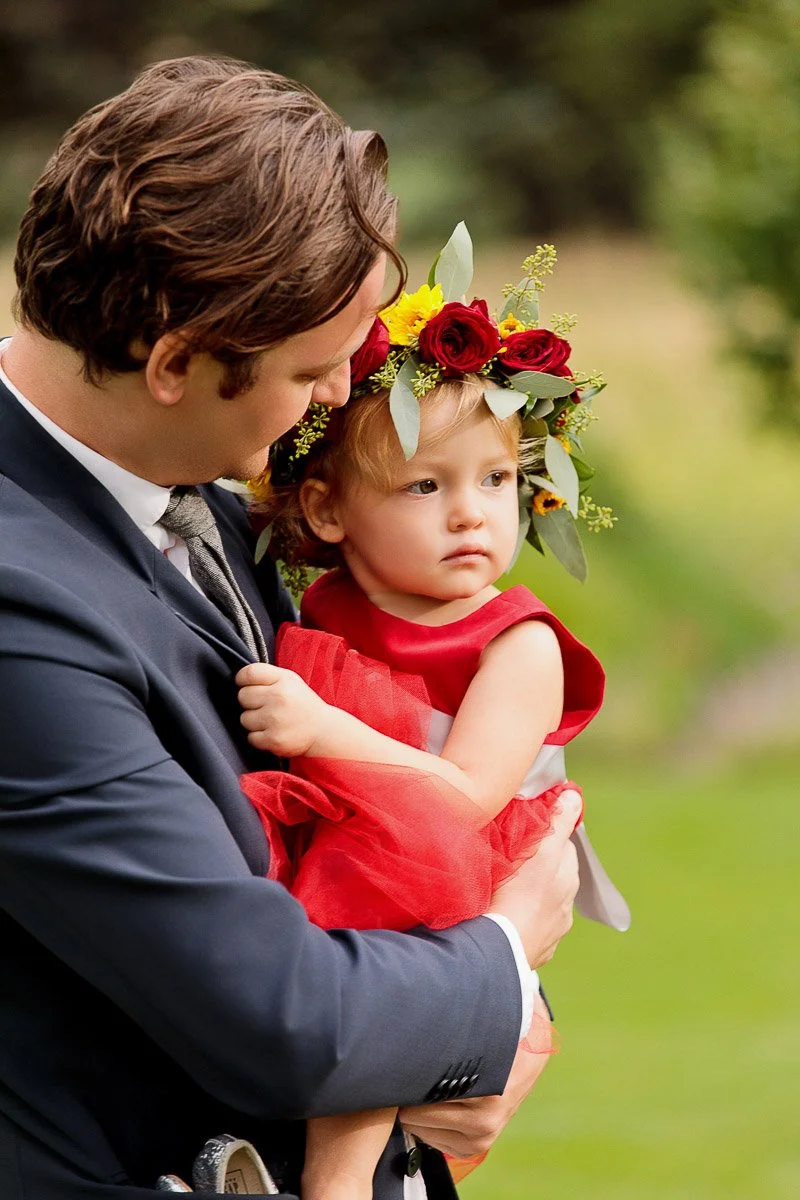 A man in a navy suit lovingly holds a young girl wearing a red dress and a floral crown, set against a blurred green outdoor background.