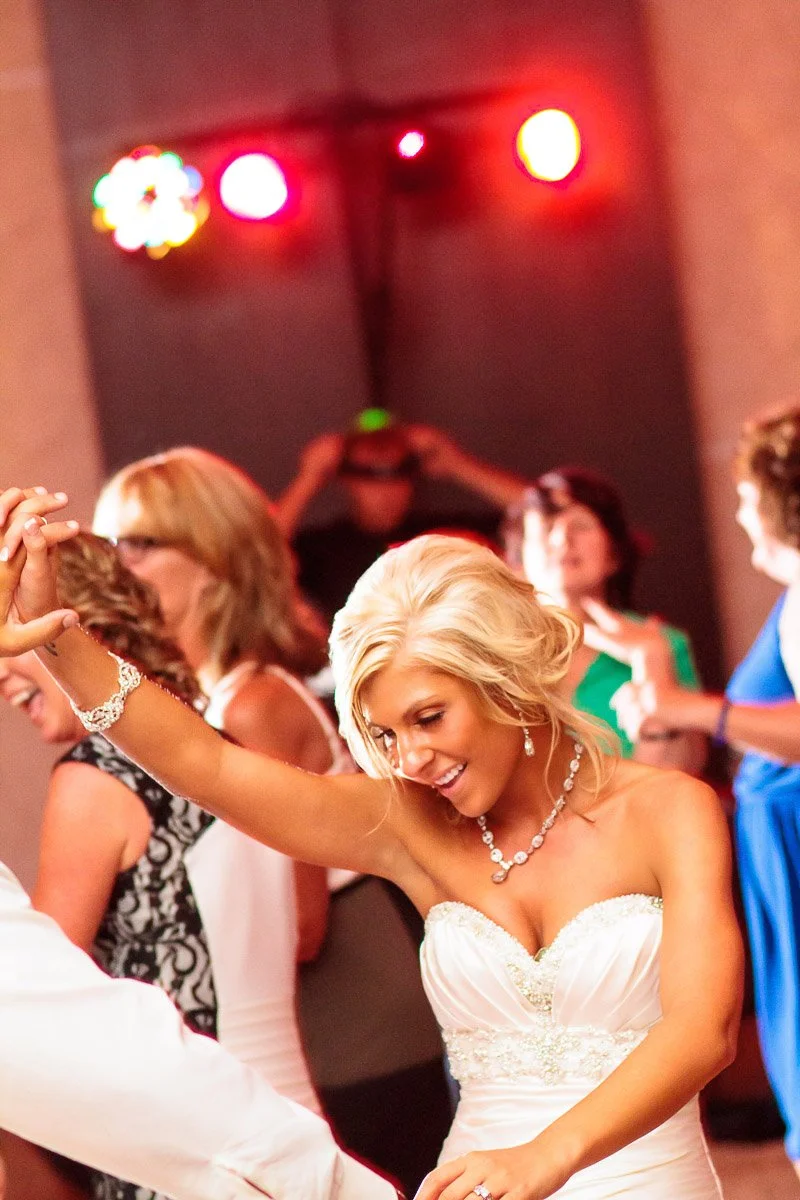 A smiling woman in a white dress dances energetically at a party. Colorful lights shine in the background, creating a joyful atmosphere.