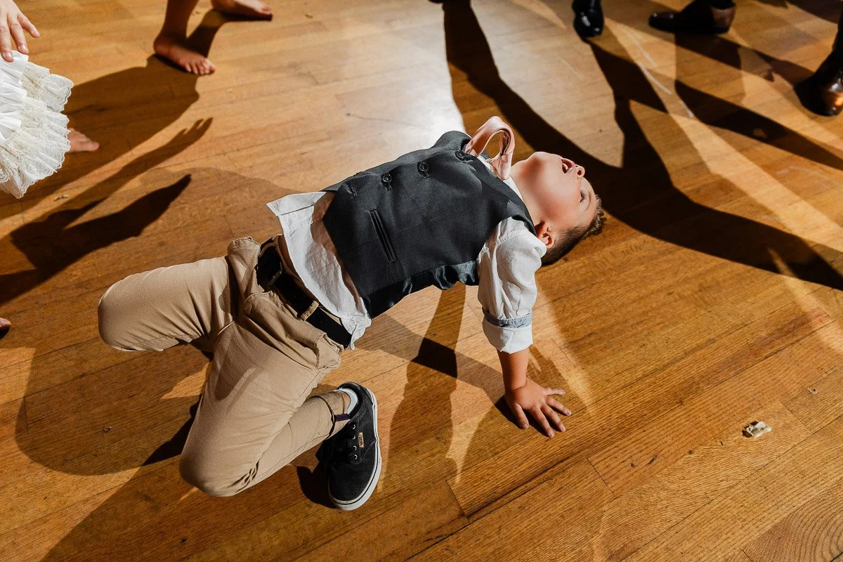 A young boy joyfully dances on a wooden floor, bending backwards. He wears a gray vest and tan pants. Surrounding shadows suggest a lively atmosphere.