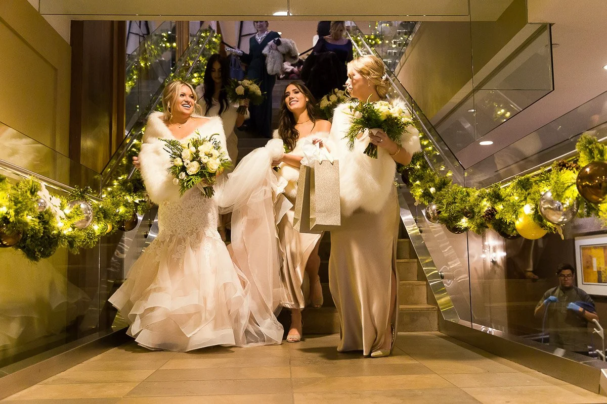 Three women in elegant dresses and fur wraps descend a festive, decorated staircase, each holding a bouquet and displaying joyful expressions.