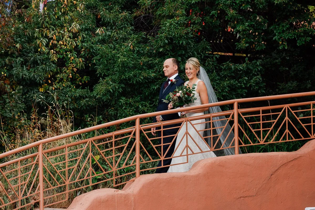 A bride and groom joyfully walk along a nature path, the bride holding a bouquet. They are surrounded by lush greenery, conveying a serene, festive atmosphere captured by Rock Rocks wedding photographer tomKphoto