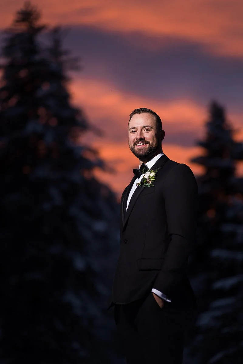 Man in a tuxedo smiling, stands confidently with hands in pockets against a backdrop of snow-covered trees and a vibrant, pink-orange sunset sky.