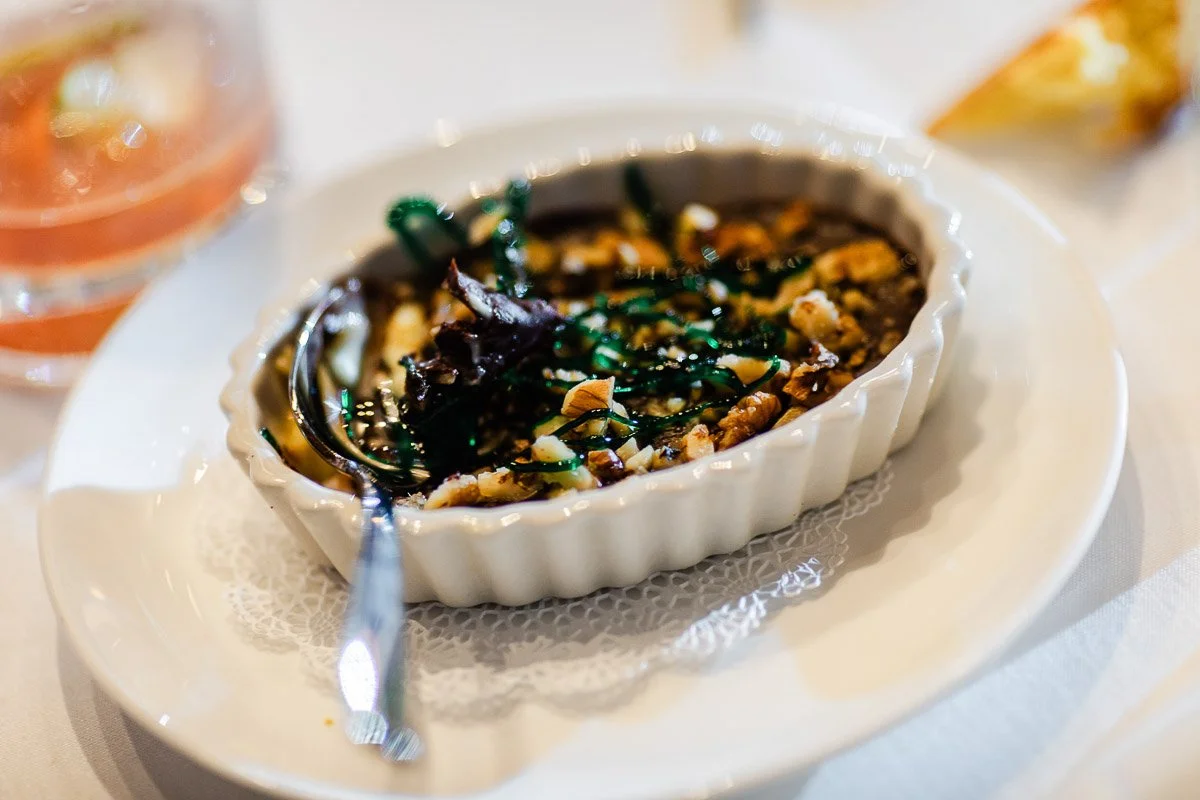 Oval dish with baked nuts and greens in a sauce, on a white doily-lined plate. A spoon is on the side, with a blurred drink in the background.