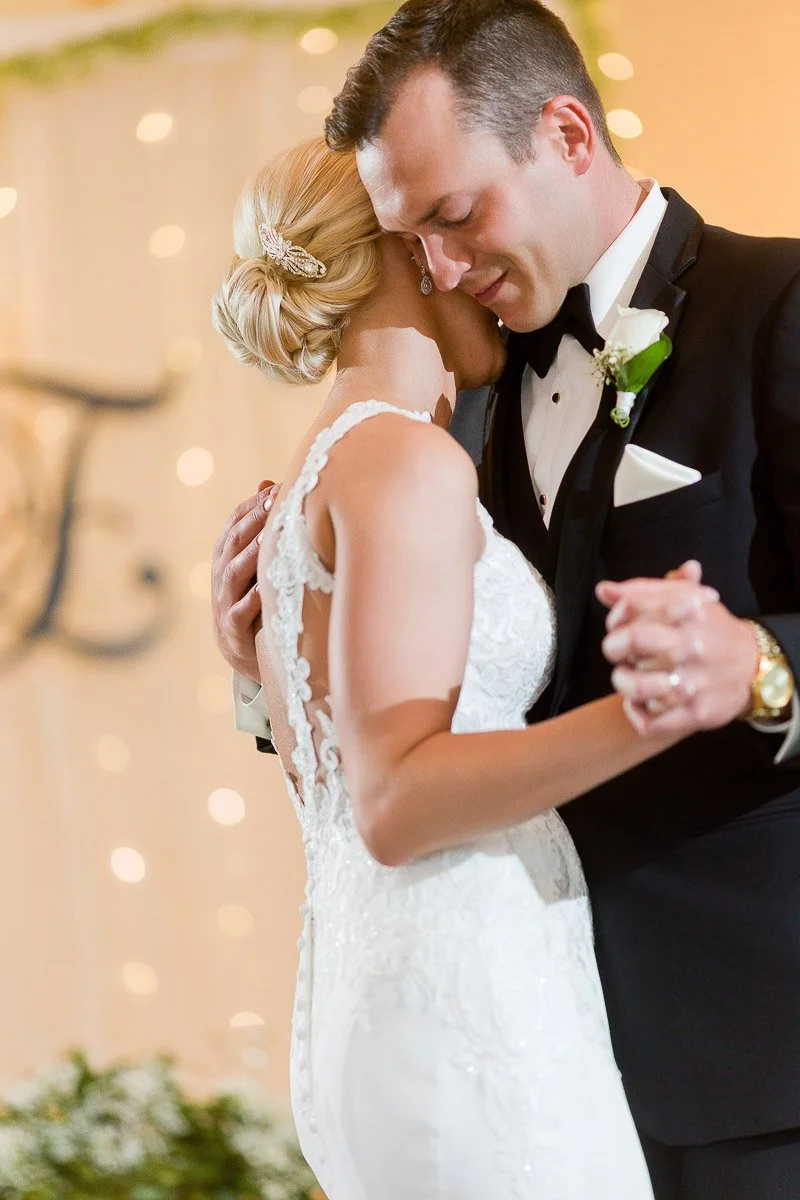A bride and groom share a tender dance, the bride in a lace gown and the groom in a tuxedo. Soft lighting creates a romantic, intimate ambiance.