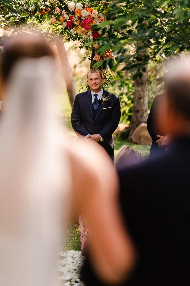A groom in a dark suit stands under a floral arch, looking emotional. Blurred foreground shows the bride, with trees and soft sunlight around.