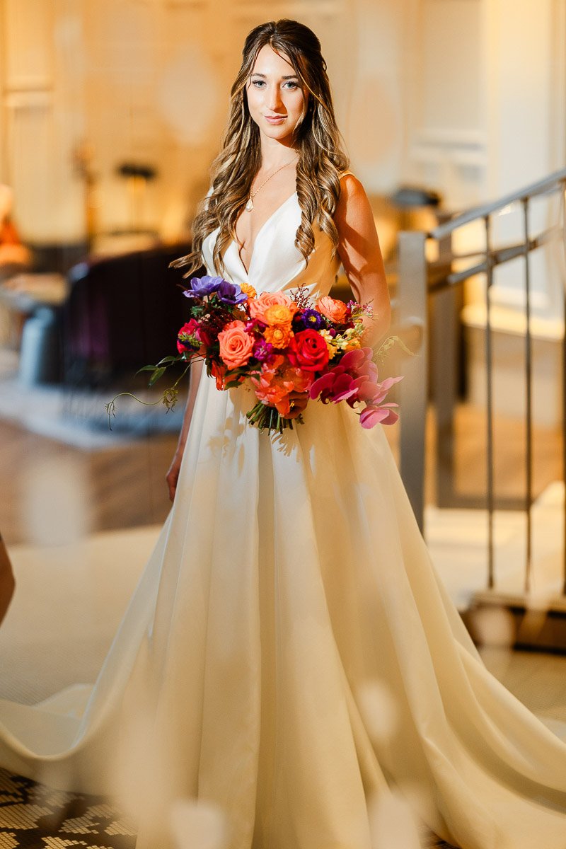 Bride in a flowing white gown holds a vibrant bouquet of red, pink, and purple flowers. Soft lighting and elegant setting evoke a serene, joyful atmosphere.