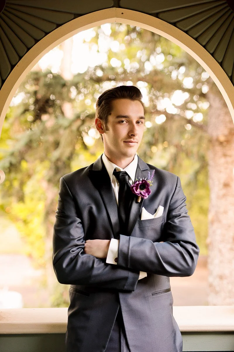 Young man in a formal grey suit with a purple boutonniere stands confidently with arms crossed under an arched window, lush greenery in the background.