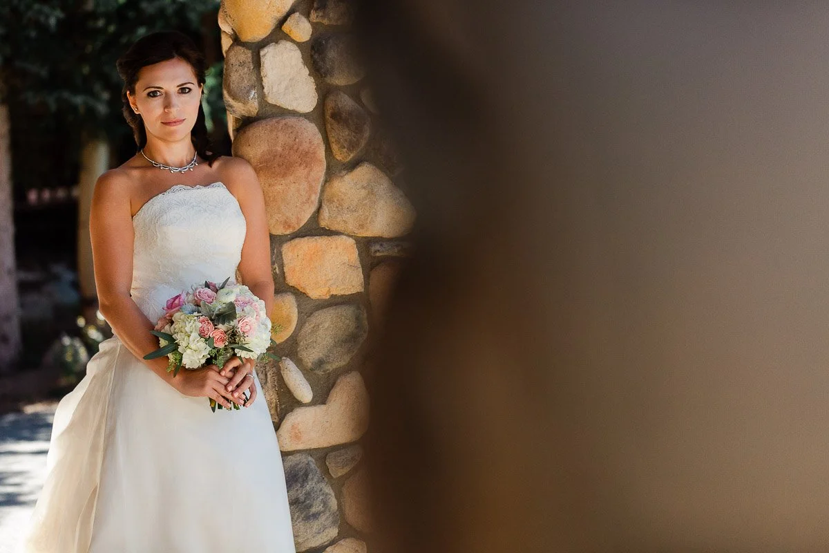 Bride in a strapless white gown holding a pastel bouquet, standing by a stone wall, looks serene and composed, with dappled sunlight on her.