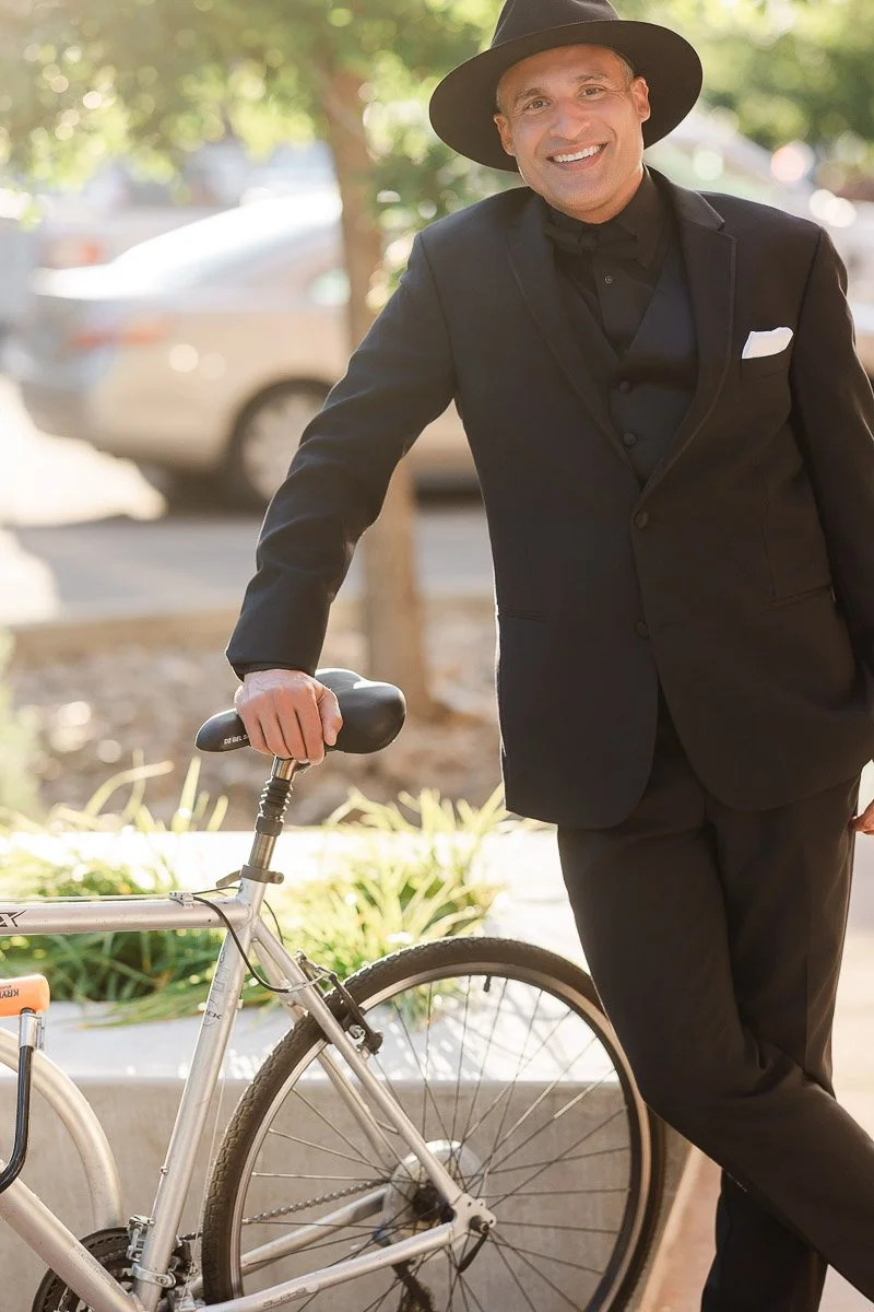 Man in a black suit and hat smiles while leaning on a bicycle. Sunlight filters through trees, casting a warm, relaxed ambiance on a city street.