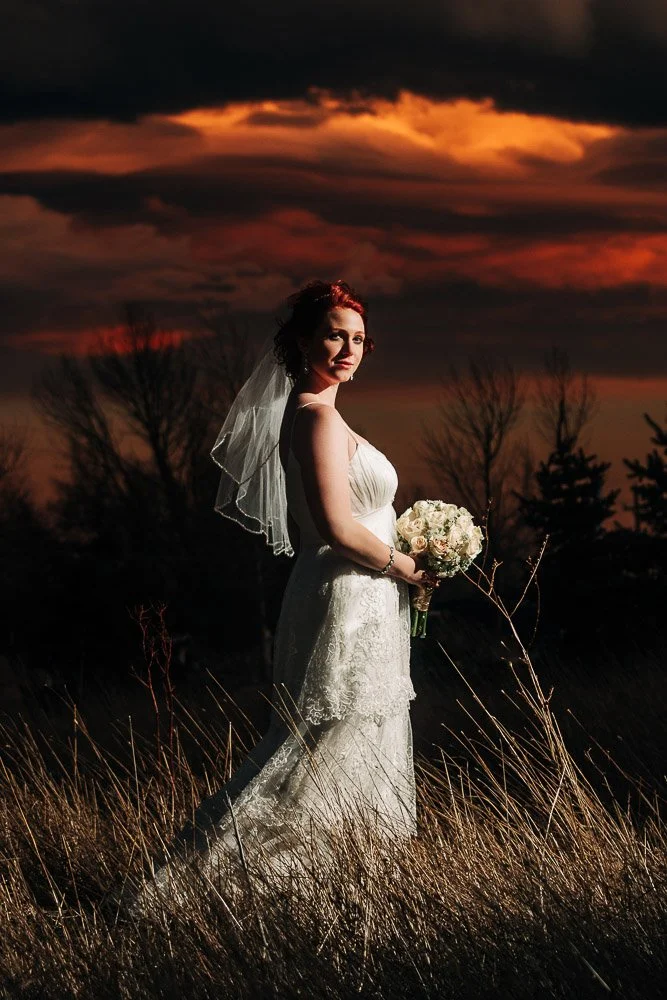 Bride in a lace gown and veil stands in a field at sunset, holding white roses. The dramatic orange sky adds a serene, romantic ambiance captured by Colorado Wedding Photographer tomKphoto