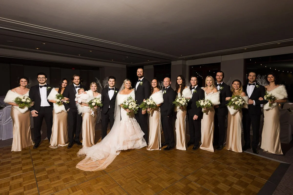 A wedding party stands elegantly, featuring a bride in a flowing dress and a groom in a tuxedo. The group, dressed in coordinated attire, holds bouquets and smiles warmly, conveying joy and celebration.