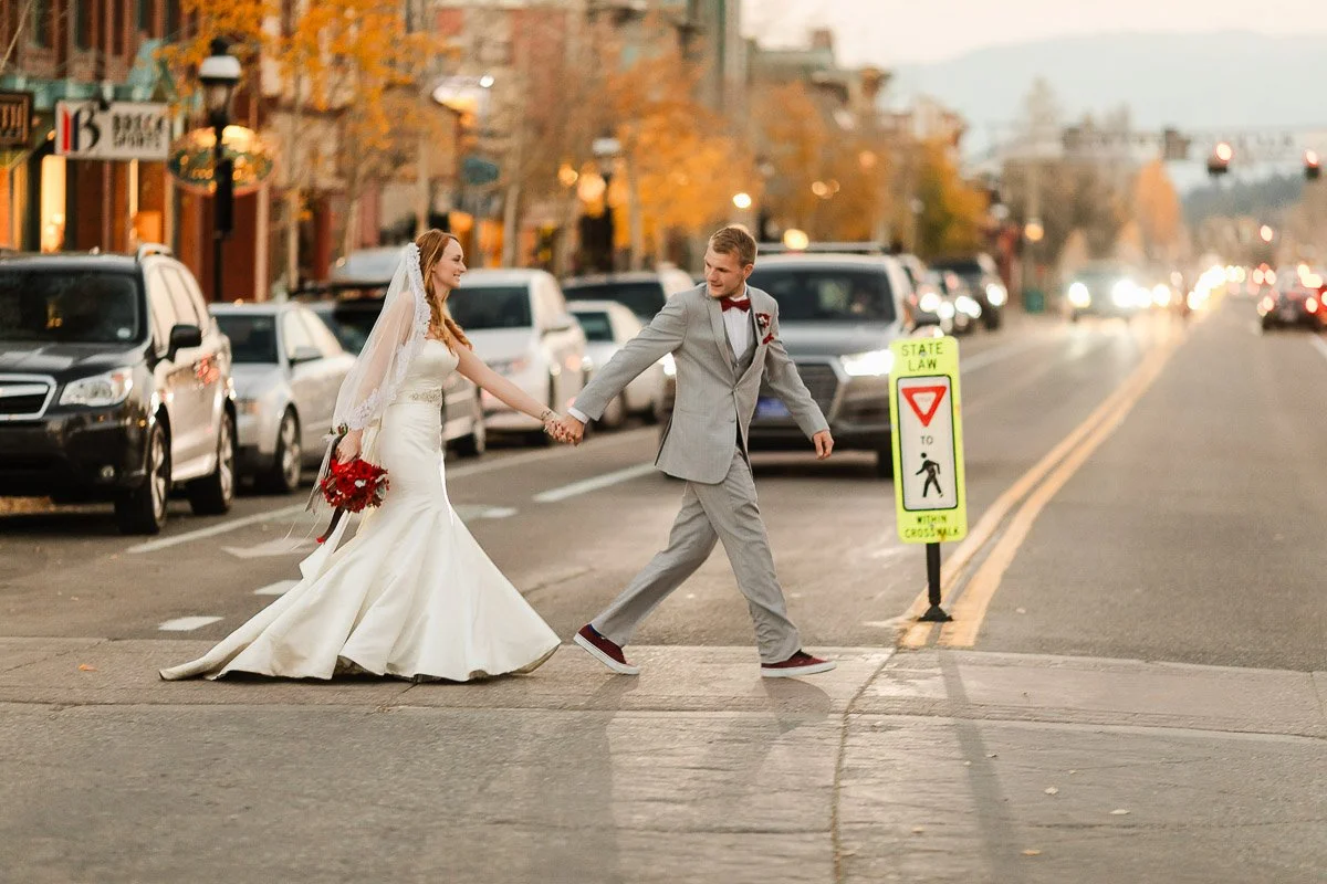 A bride and groom hold hands, crossing a city street at sunset. She wears a white gown, he a gray suit, as cars line the road, creating a romantic scene.
